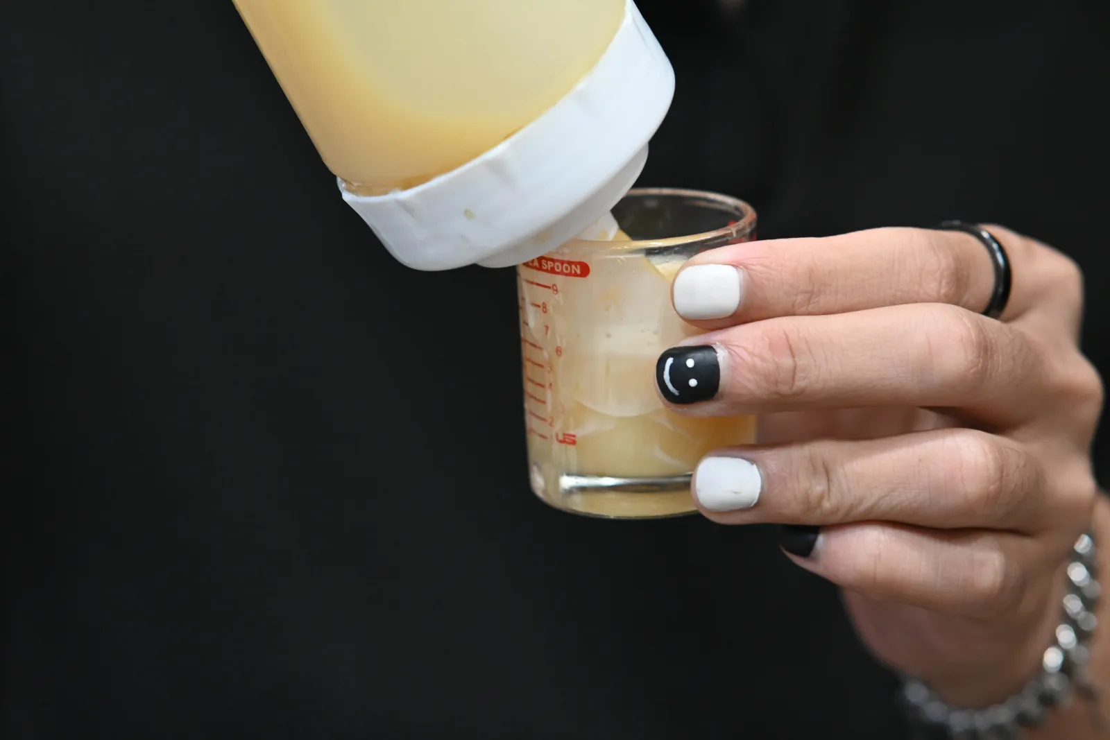 This close-up photograph captures a hand with striking black-and-white manicured nails holding a small glass measuring cup, into which a pale yellow liquid is being poured from a white squeeze bottle. The nails are predominantly painted glossy white, with one accent nail featuring a black design, and a silver ring adorns one of the fingers. The measuring glass, marked with red measurement lines, is partially filled with the translucent yellow liquid. The dark, out-of-focus background - likely black clothing - creates strong contrast that draws attention to the hand, the nail art, and the careful pouring action taking place, suggesting a moment from a cocktail preparation or similar mixing process, fitting for the setting of Pattaya City, Thailand.