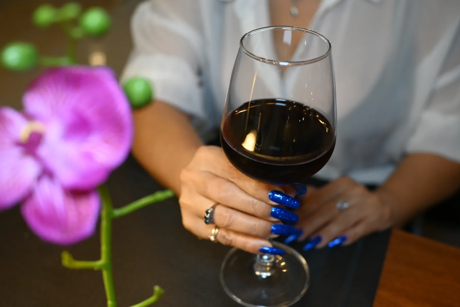 This photo shows someone holding a glass of red wine at what appears to be a restaurant or dining establishment. The person is wearing a light-colored (white or light blue) shirt and has striking blue nail polish on their manicured nails, along with a ring. In the foreground, there's a beautiful purple orchid flower that adds an elegant touch to the scene. The setting appears to be indoors with ambient lighting creating a warm, intimate atmosphere. The photo seems to capture a moment during a meal or social gathering, with the wine glass being the focal point of the image.