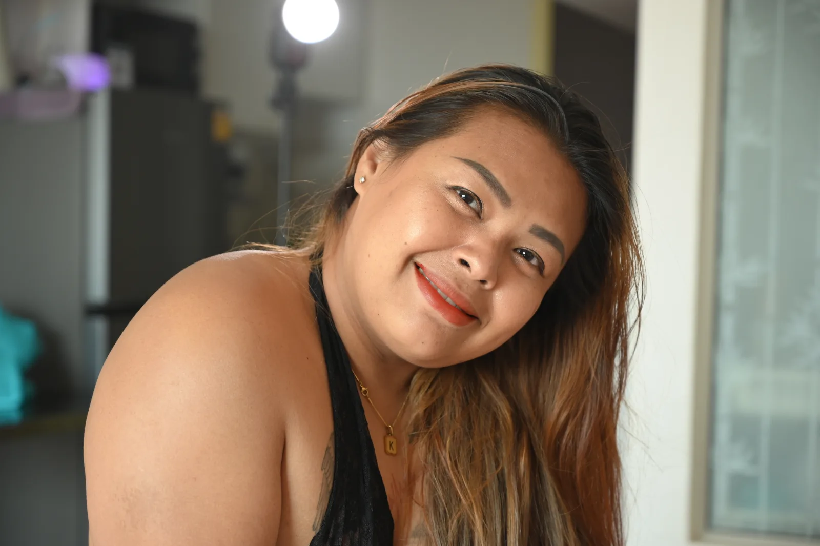 This photo features a beautiful young woman with long, dark hair and a bright smile. She is wearing a black top and a gold necklace, which adds a touch of elegance to her appearance. The woman is posing for the camera, and her smile radiates warmth and happiness. The scene appears to be set in a room with a chair visible in the background.