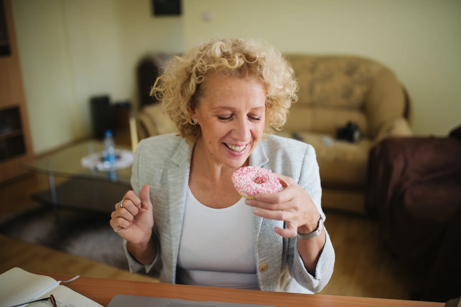 Elderly woman with curly blonde hair smiling while eating a donut with pink frosting