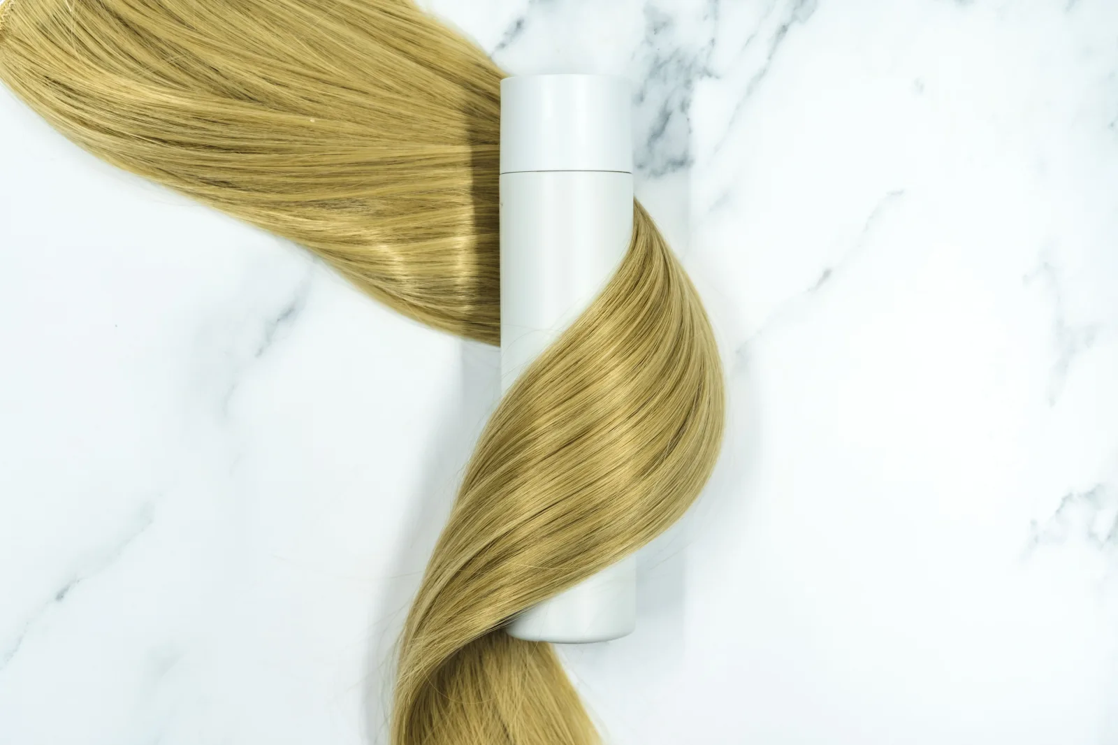 In this image, a lock of long blonde hair can be seen alongside a bottle of hair treatment or product on a white marble background. We originally took this photo for use on our own website for a blogpost about hairstyling products, but have now decided to release it under Creative Commons Attribution Licensing. This means we're more than happy for you to use it in any way you wish (woohoo!) But please be sure to credit us when you do, by including a link to geehair.com/