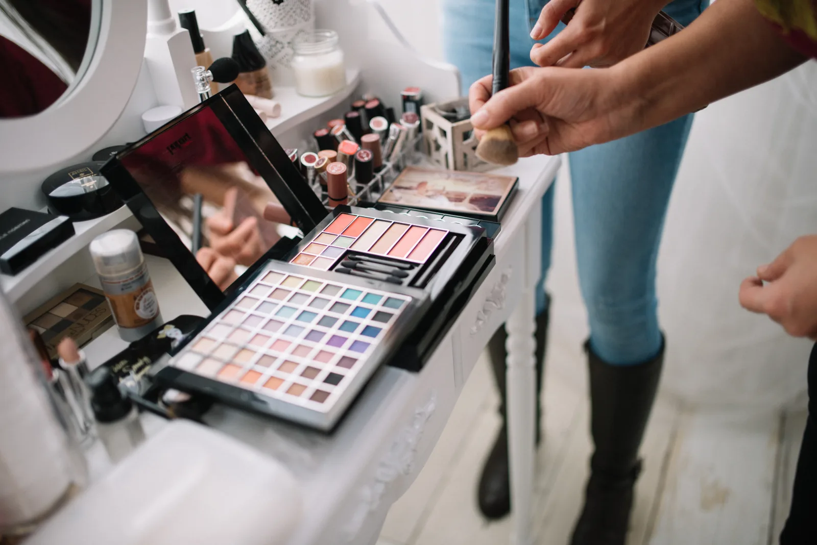 Hands of a makeup artist are shown holding a brush while colorful makeup palettes are arranged neatly on a clean table in a well-lit studio, highlighting creativity and artistry.