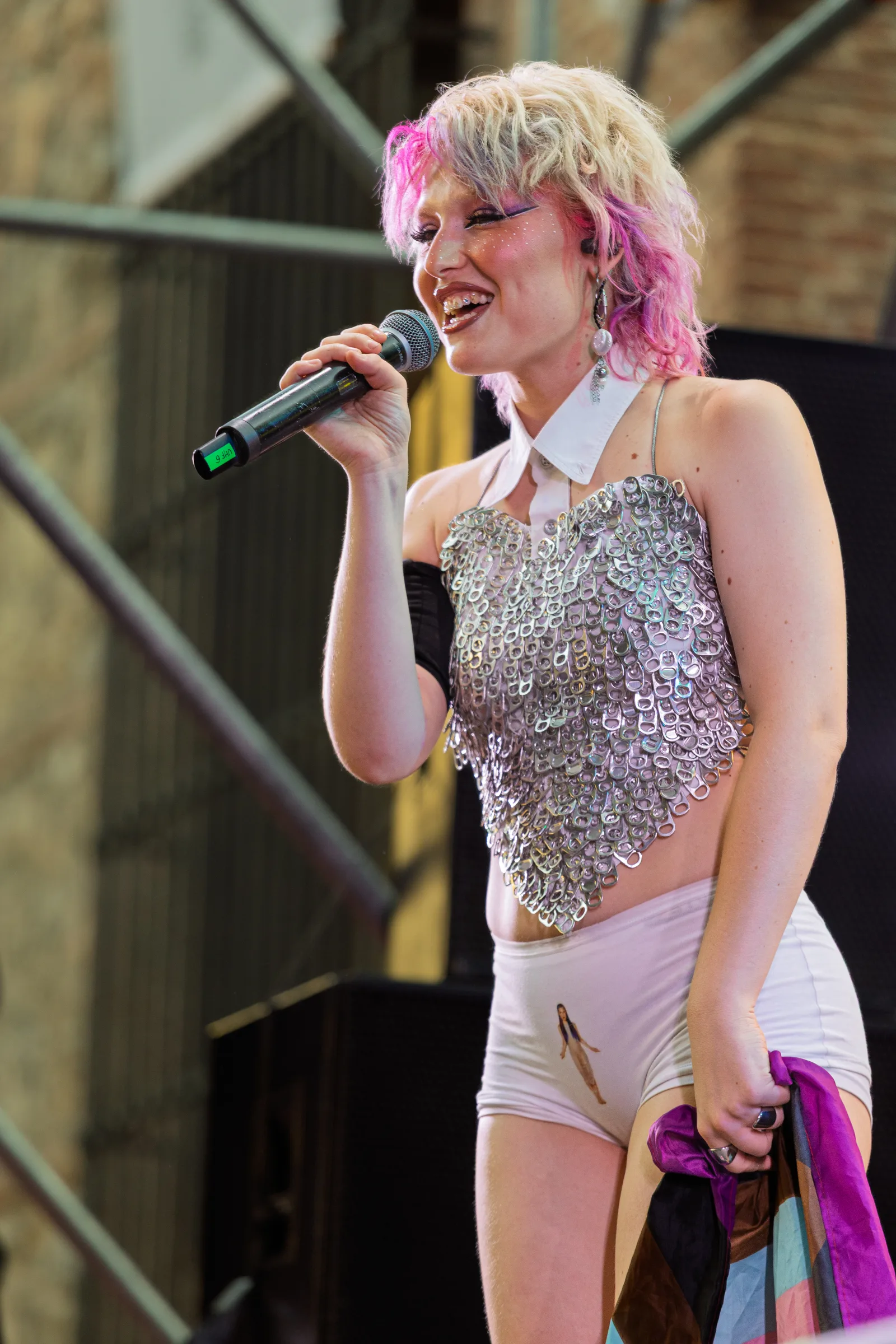 Spanish singer Sofía Coll addresses the audience at Plaza del Rey during the opening moments of her performance at the Madrid Pride celebrations on July 4, 2025. Dressed in a sparkly sequined crop top and light-colored shorts, with her platinum blonde hair accented by subtle pink highlights, Coll appears composed and confident as she connects with the crowd before launching into her high-energy set. The warm stage lighting adds to the welcoming atmosphere of this early part of her Pride performance.