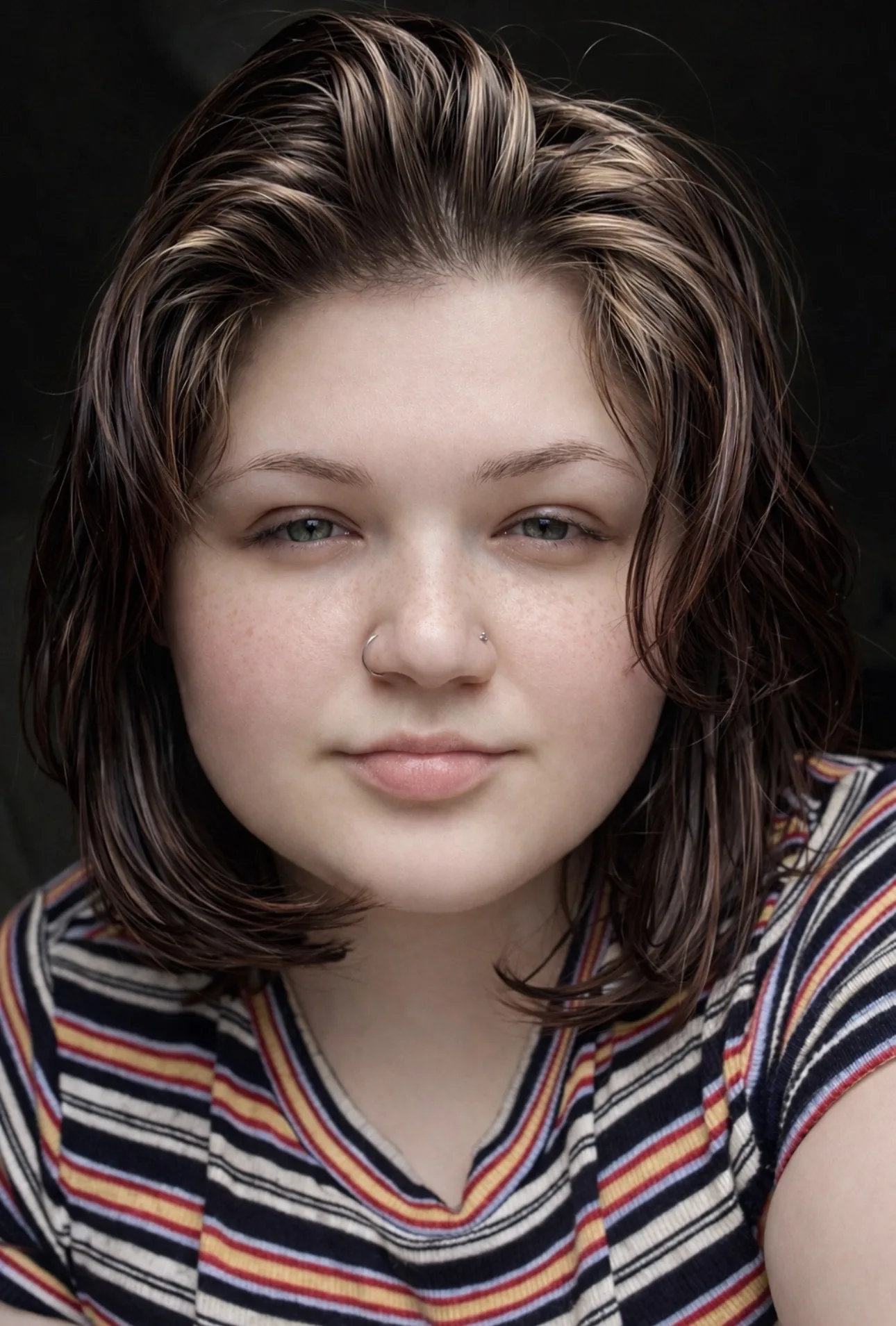 This is a close-up portrait of Sidney Lynn with a really natural, indie-style aesthetic. She’s facing the camera directly with a calm, slightly soft expression that feels very genuine and unposed.
Her hair is short to medium length, tousled, and softly layered with warm brown tones and subtle highlights, giving it that effortless, slightly messy look that fits the indie vibe. Her skin shows natural texture and light freckles across her cheeks and nose, which adds to the authenticity instead of a heavily edited feel. She has a small nose ring and an additional subtle piercing, which gives her look a bit of an alternative edge.

She’s wearing a striped shirt with warm tones, and the background is dark and blurred, which keeps the focus entirely on her face. The lighting is soft and even, highlighting her features gently without harsh contrast.