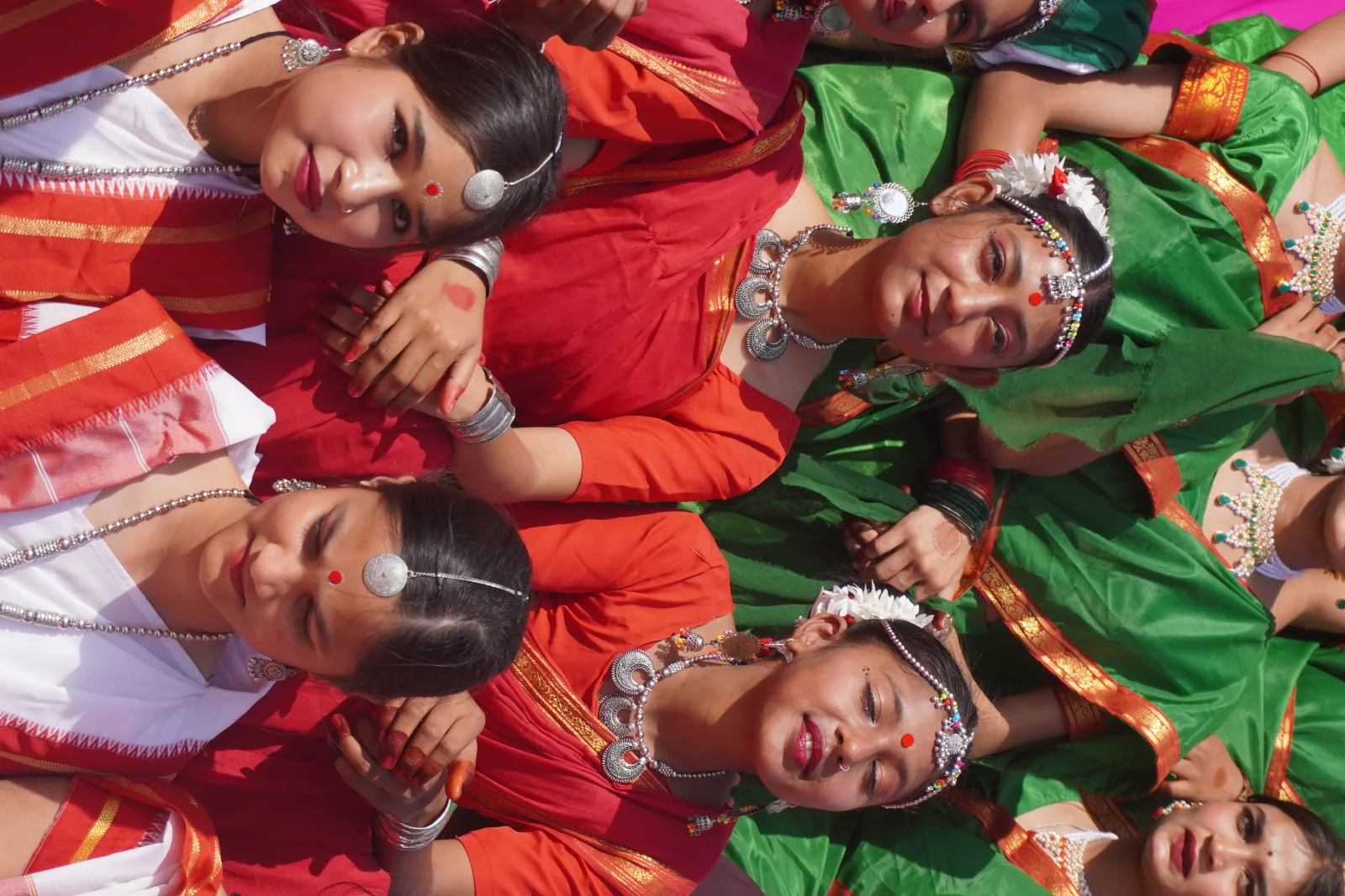 a group of women in traditional Indian sarees, predominantly in red, white, and green, participating in a cultural or folk festival. Their attire, jewelry, and makeup suggest a connection to tribal or regional dance forms from eastern India, such as the Santal or Bihu dance. The red bindis, silver jewelry, and floral hair adornments add to their graceful and elegant appearance. The expressions on their faces, ranging from calm to joyful, indicate a moment of unity and cultural pride as they prepare for their performance under the bright sunlight.