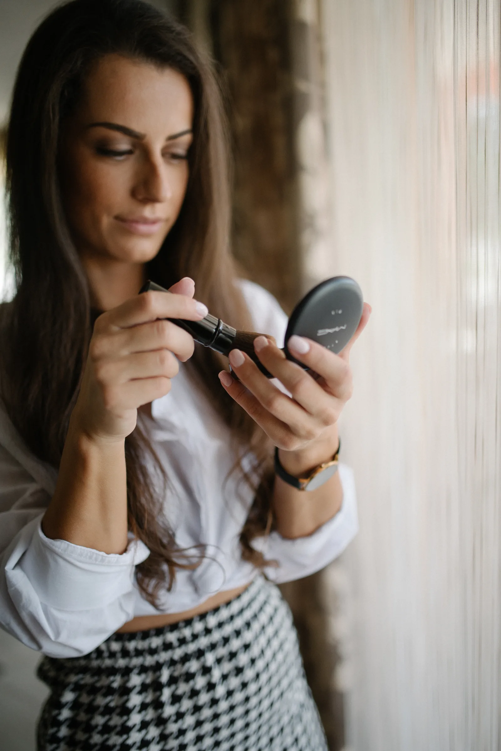 A woman with long hair focuses on applying makeup while holding a mirror. She wears a fashionable outfit and stands by a window, illuminated by natural light.