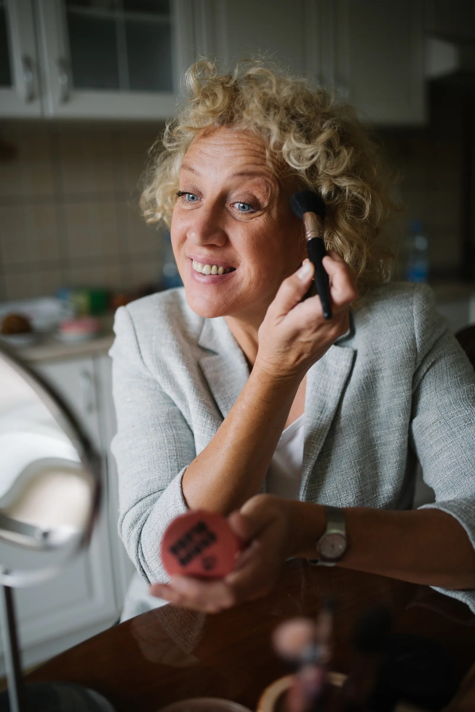 Woman with blonde curly hair putting on makeup powder and smiling