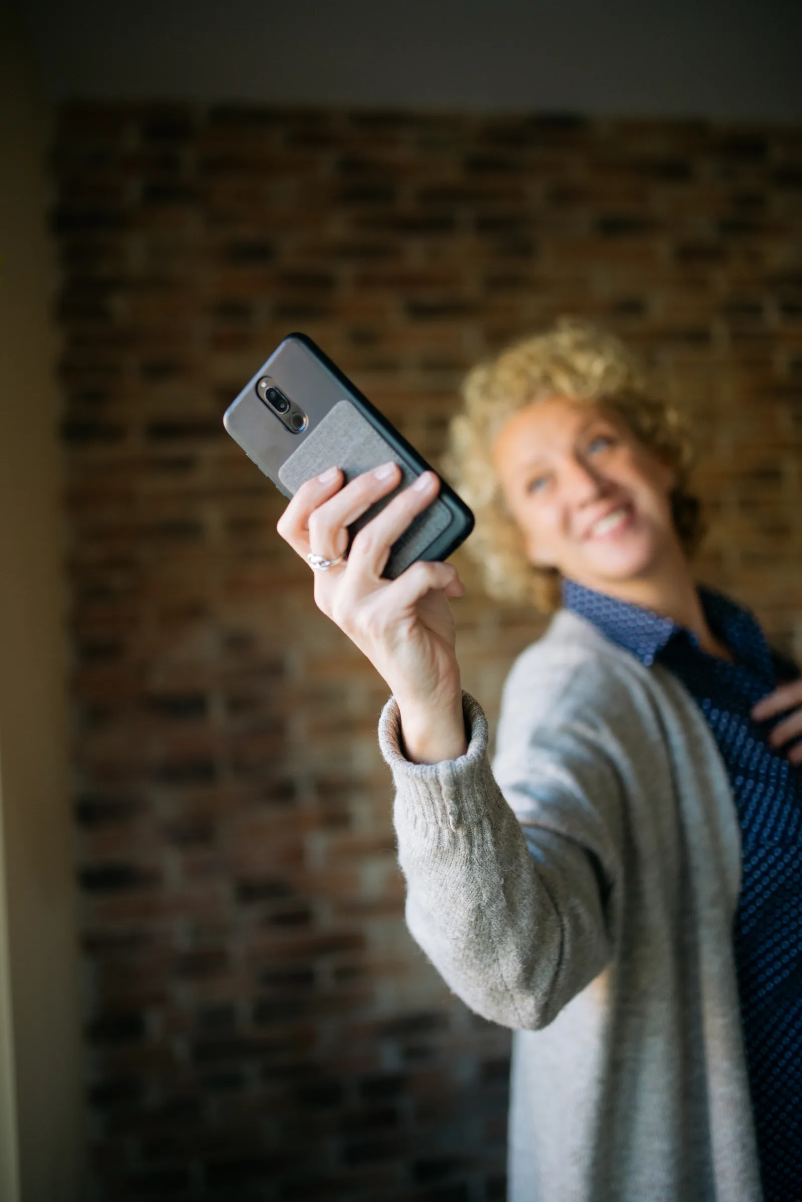A woman with curly blonde hair happily takes a selfie in a comfortable indoor space. The warm atmosphere and brick wall create a relaxed backdrop for her cheerful expression.