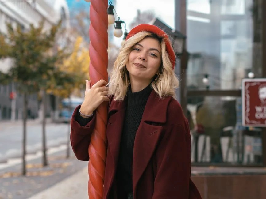 Chic woman smiling in red beret and coat, leaning on a column in a vibrant city street.