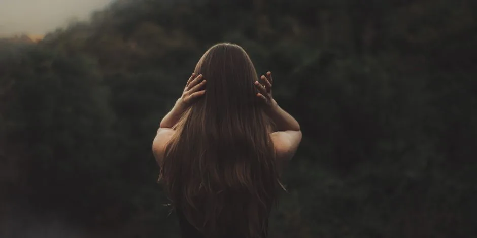 A woman with long hair stands in a forest, backlit by soft light, with her hands in her hair.