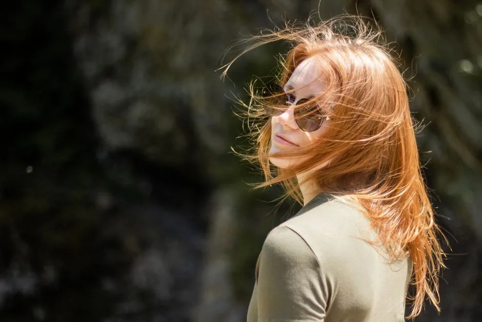 A smiling woman with red hair enjoying a sunny day wearing sunglasses.