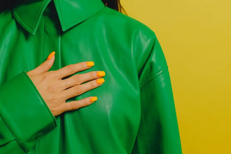 Close-up of a woman's hand with yellow nails on a green leather jacket against a yellow background.