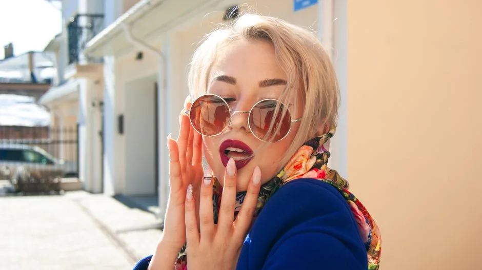 Trendy blond woman with sunglasses and floral scarf posing outdoors, showcasing chic fashion and manicured nails.