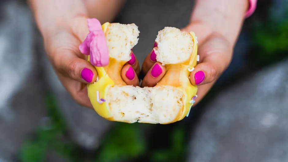 A vibrant image showing pink manicured hands gently tearing a yellow-frosted doughnut in half.