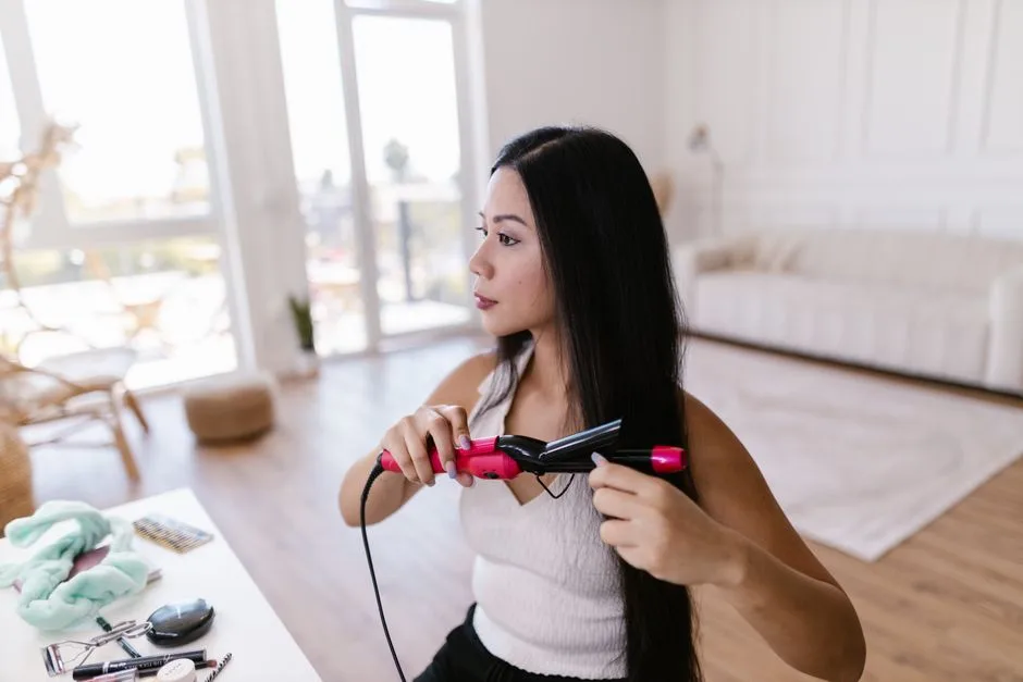 Asian woman using a flat iron at home, achieving a sleek hairstyle in a modern interior setting.