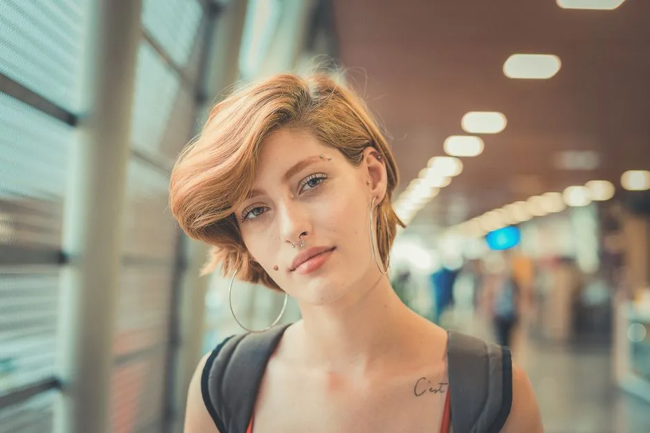 A confident young woman with short hair and piercings standing in a modern indoor setting.