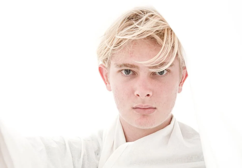 Striking close-up of a blond young man with wind-swept hair against a bright backdrop.