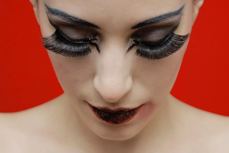 Close-up of a woman with striking makeup and dramatic eyelashes against a red background.
