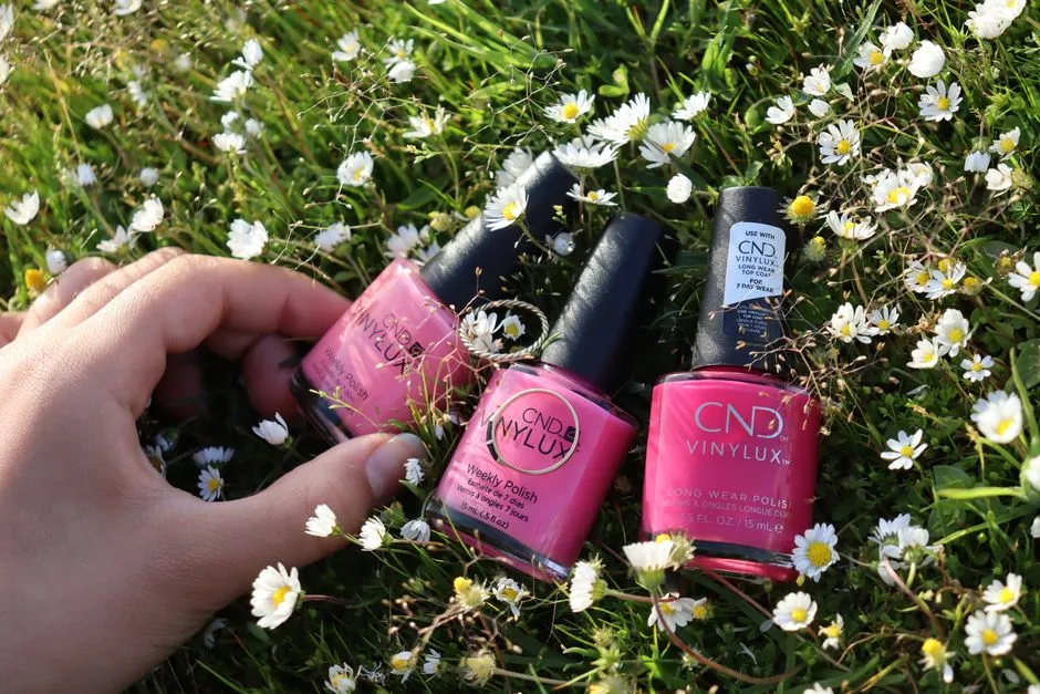 Vibrant pink nail polishes displayed on grass surrounded by daisies.