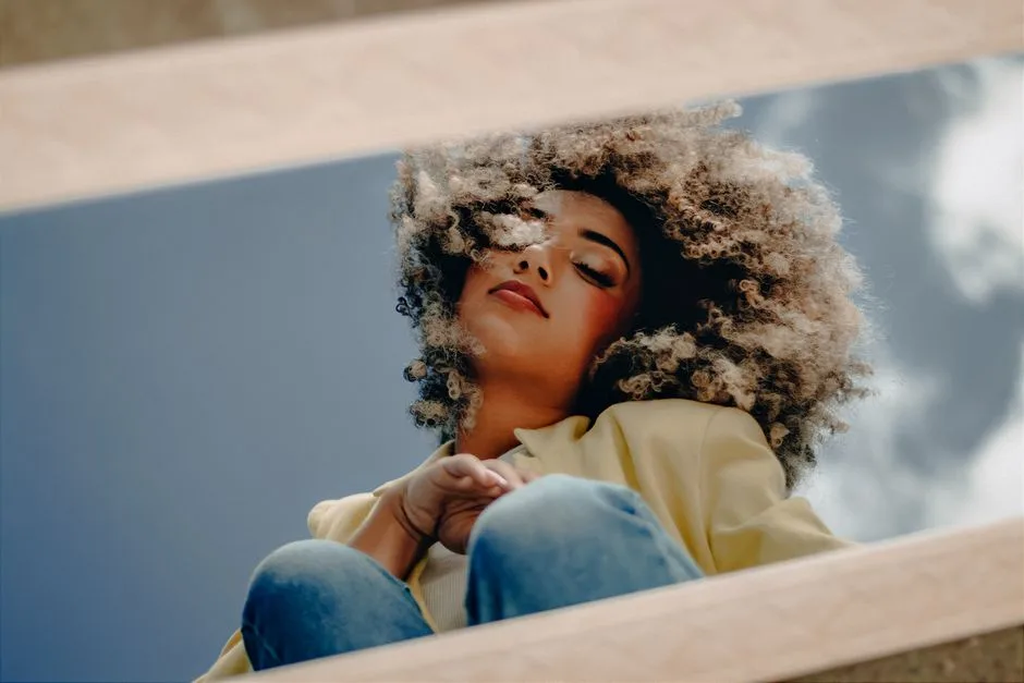Woman with curly afro hair in a yellow jacket peacefully posing outdoors against a bright blue sky.