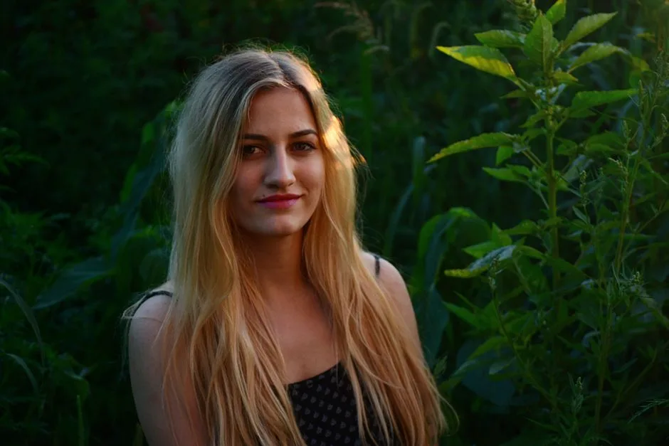 A beautiful blonde woman smiling in a natural outdoor setting with lush greenery.