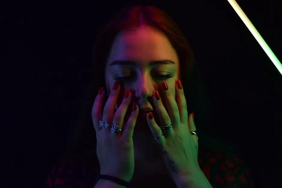 Close-up portrait of a woman with colorful neon lights and closed eyes, showcasing rings and red nails.
