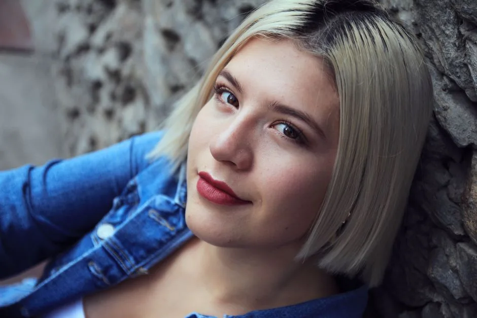 Close-up portrait of a young woman with blonde hair in a denim jacket smiling against a textured background.