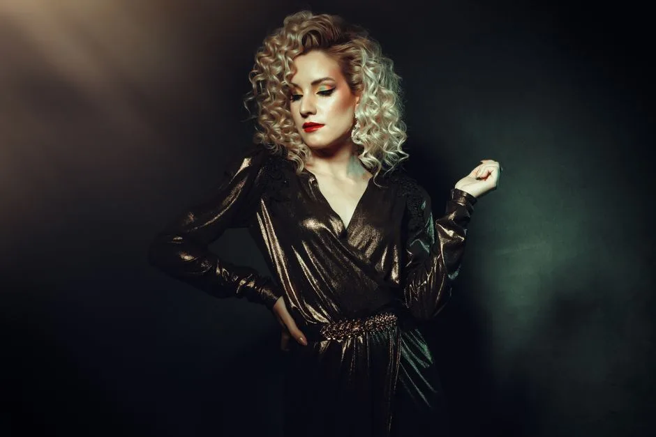 Stylish woman with curly blond hair posing in a shimmering outfit against a dark studio backdrop.