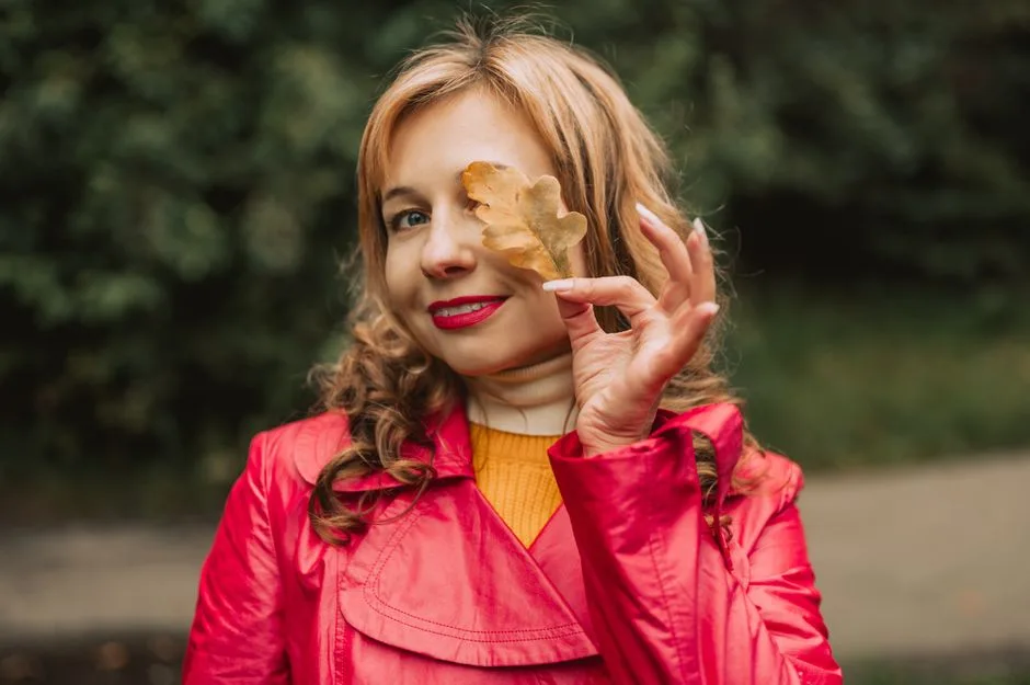 A smiling woman in a red coat playfully holds an oak leaf in an autumn setting.