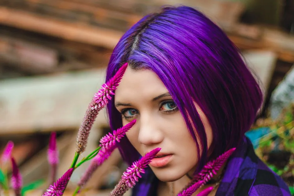Close-up portrait of a woman with purple hair among vibrant flowers.
