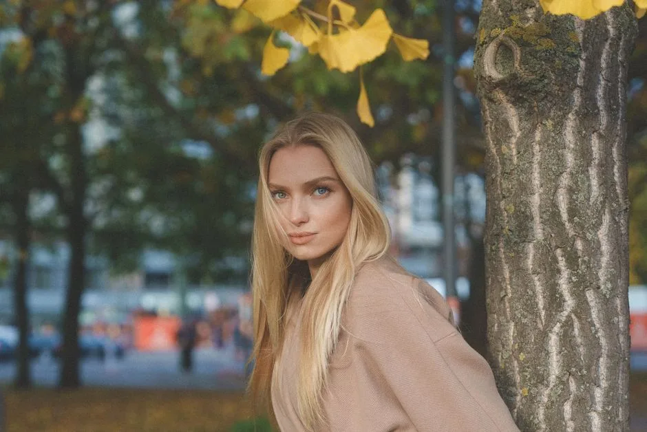 A fashionable woman posing by a tree with golden ginkgo leaves in a park setting.