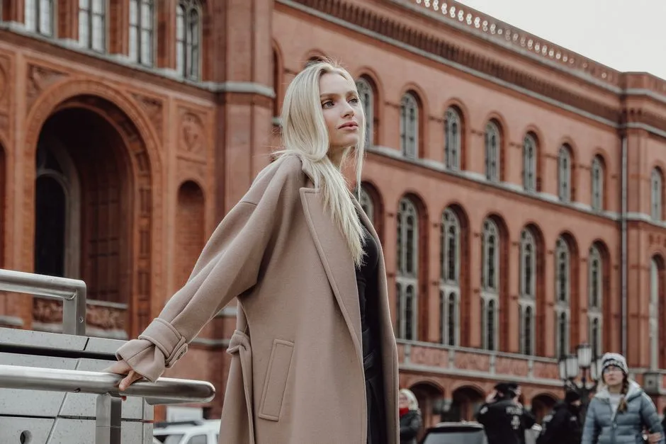 Stylish woman with long blonde hair poses elegantly in front of a historic red brick building in an urban setting.