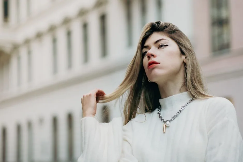 Portrait of a stylish woman posing outdoors with a chain necklace and white sweater.