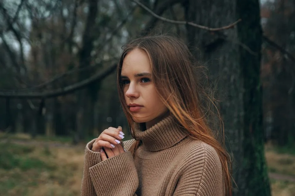 A contemplative woman poses in a woodland setting, wearing a cozy brown sweater on a fall day.
