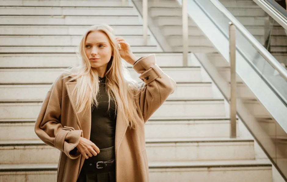 Stylish woman in a beige coat posing on modern city stairs, exuding urban elegance.