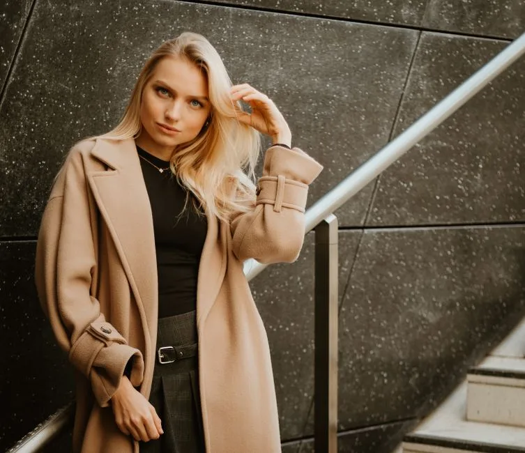 Fashionable woman in a beige coat posing on a modern staircase in an urban setting.
