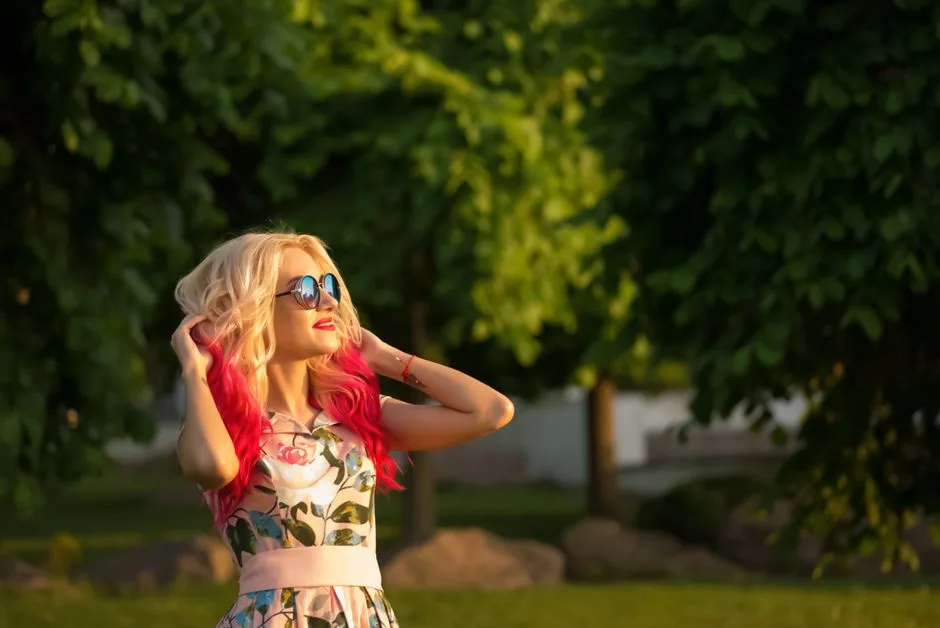 A stylish woman with colorful hair enjoys a sunny day in a park, capturing the joy of summer.