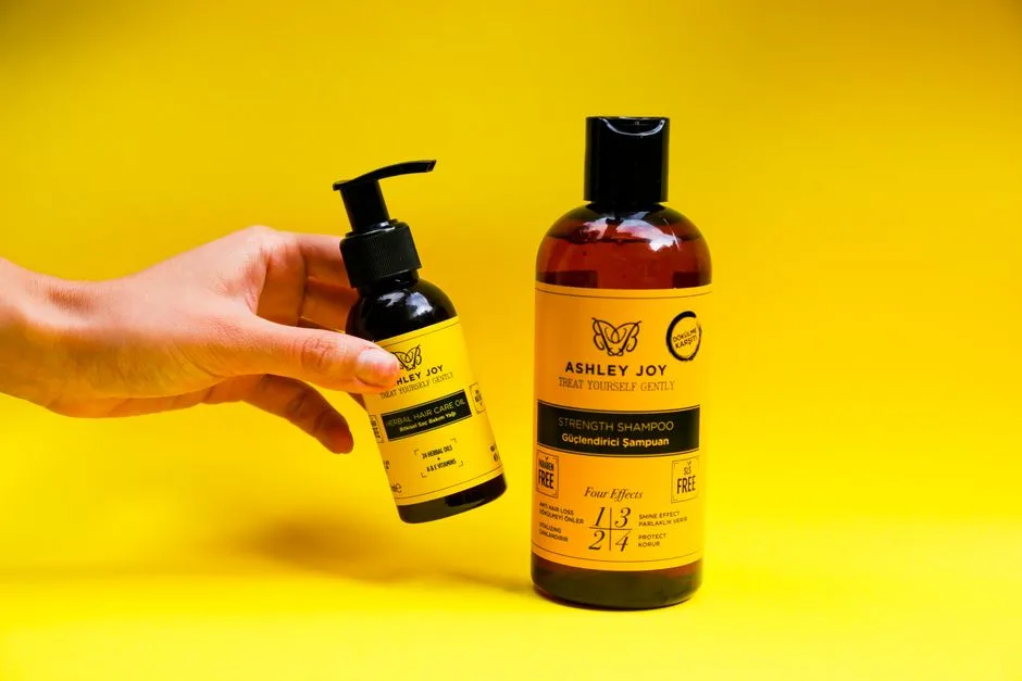 Hand holding a bottle of herbal hair care oil next to shampoo on yellow backdrop.