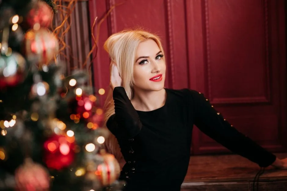 Blonde woman in black dress posing indoors next to decorated Christmas tree with red ornaments.