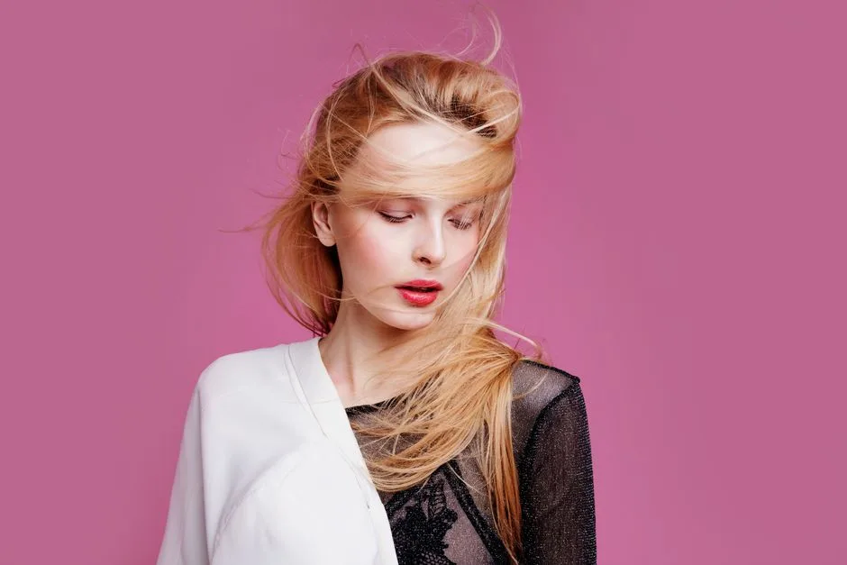 Fashion portrait of a blonde woman with hair blowing gracefully against a pink backdrop, exuding elegance.