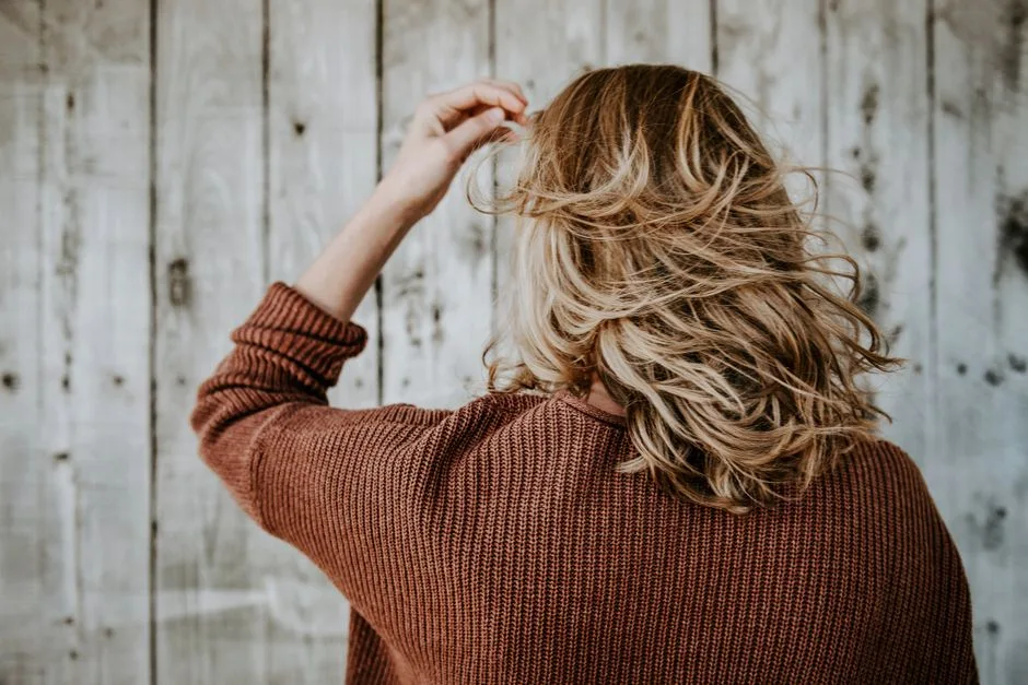 Rear view of a woman with blonde hair wearing a cozy sweater in front of rustic wooden wall.