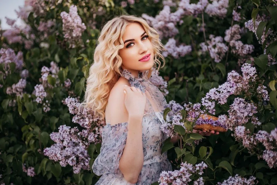 Stylish blonde woman in a sundress surrounded by lilac flowers. Perfect fashion portrait.