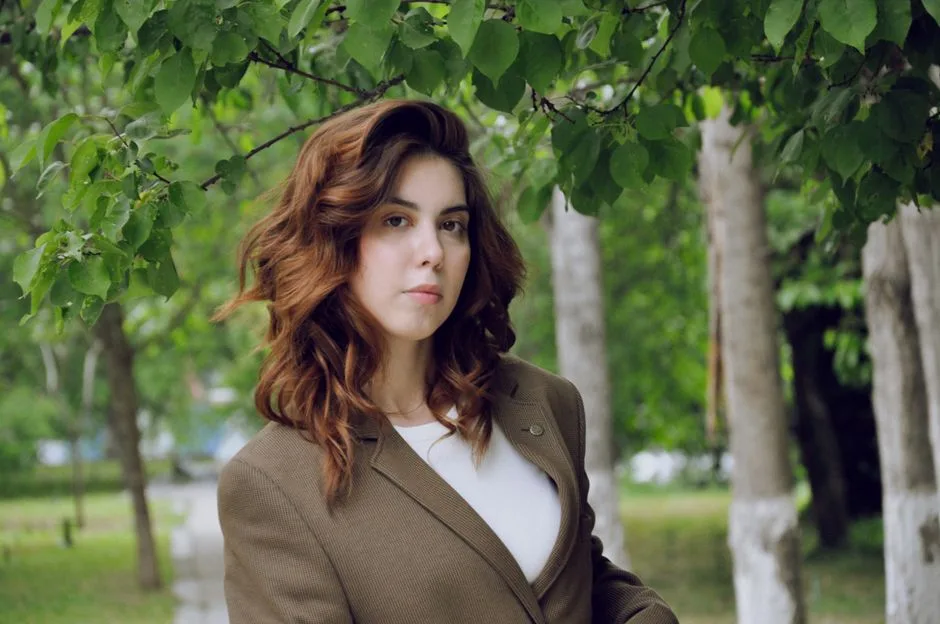 Woman with wavy hair wearing a brown blazer, standing confidently in a lush green outdoor setting.