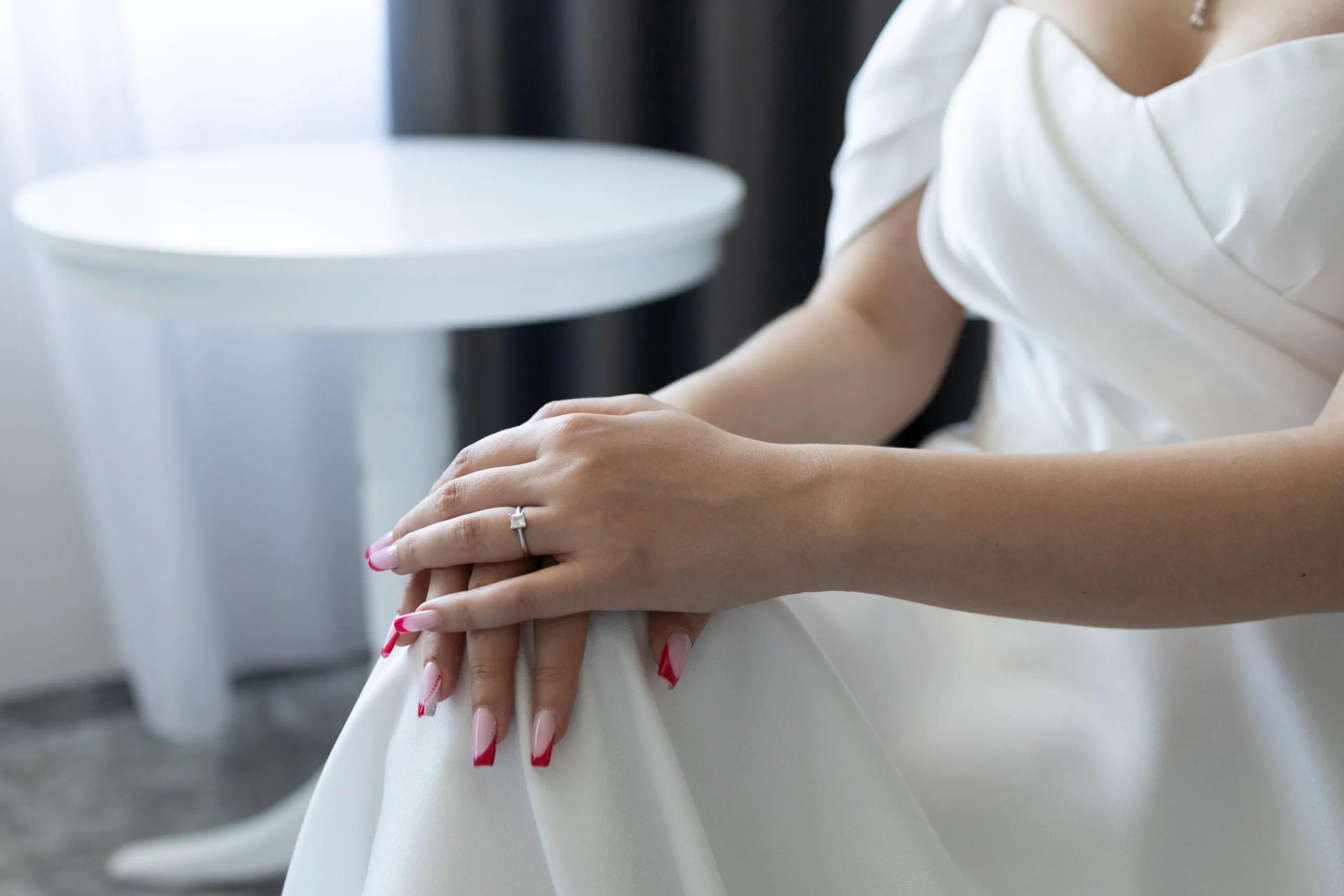 Bride in a white dress displaying her engagement ring with elegant pink nails indoors.
