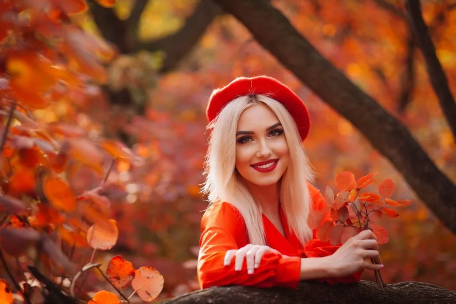 Blonde woman in red attire smiling among fall foliage, exuding warmth.