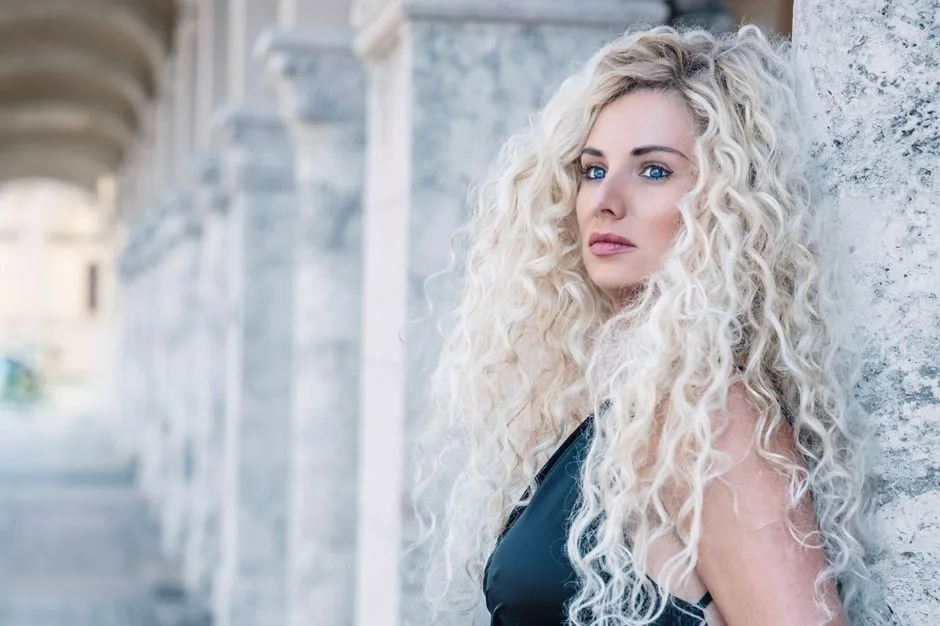 Stylish outdoor portrait of a woman with curly blonde hair leaning against a stone column.