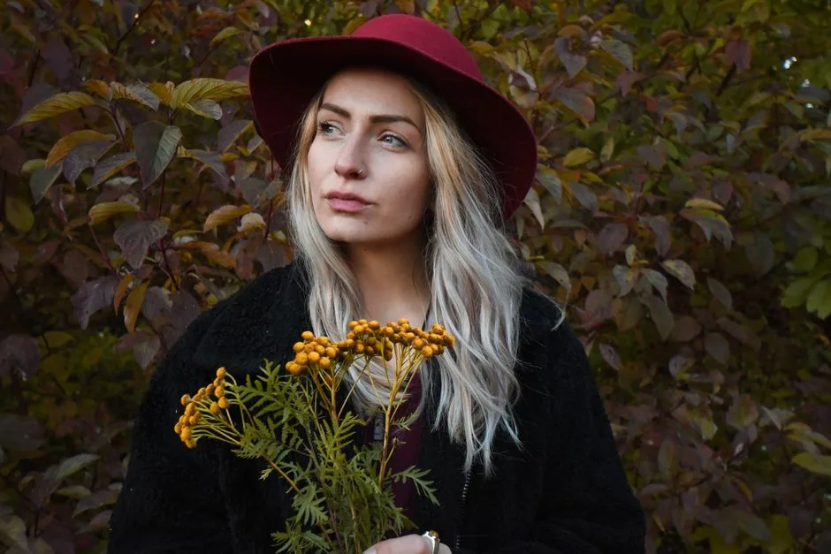 Fashionable woman with blonde hair and red hat holding yellow flowers in an outdoor setting filled with greenery.