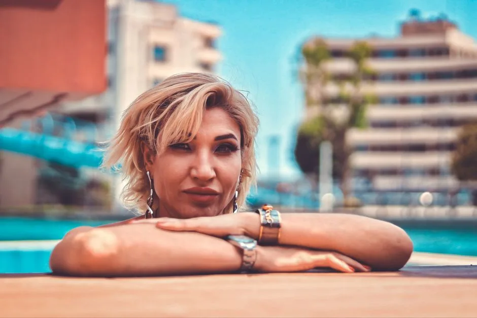 Blonde woman leaning on pool edge with cityscape in background, relaxed mood.