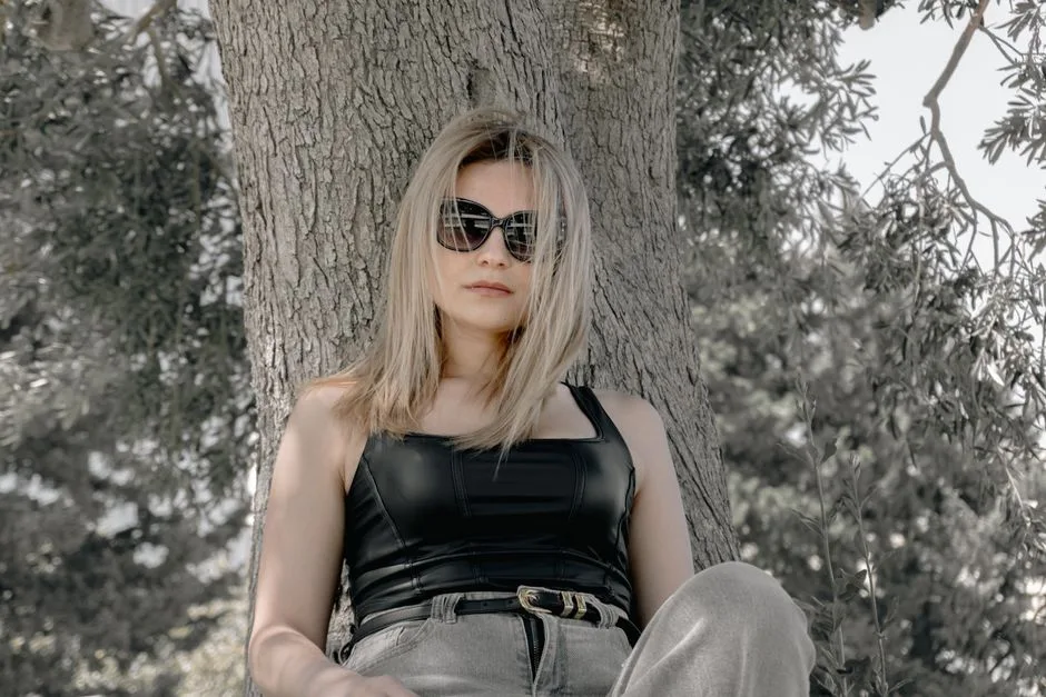 Blonde woman wearing sunglasses and casual outfit relaxing against a tree enjoying a sunny day outdoors.