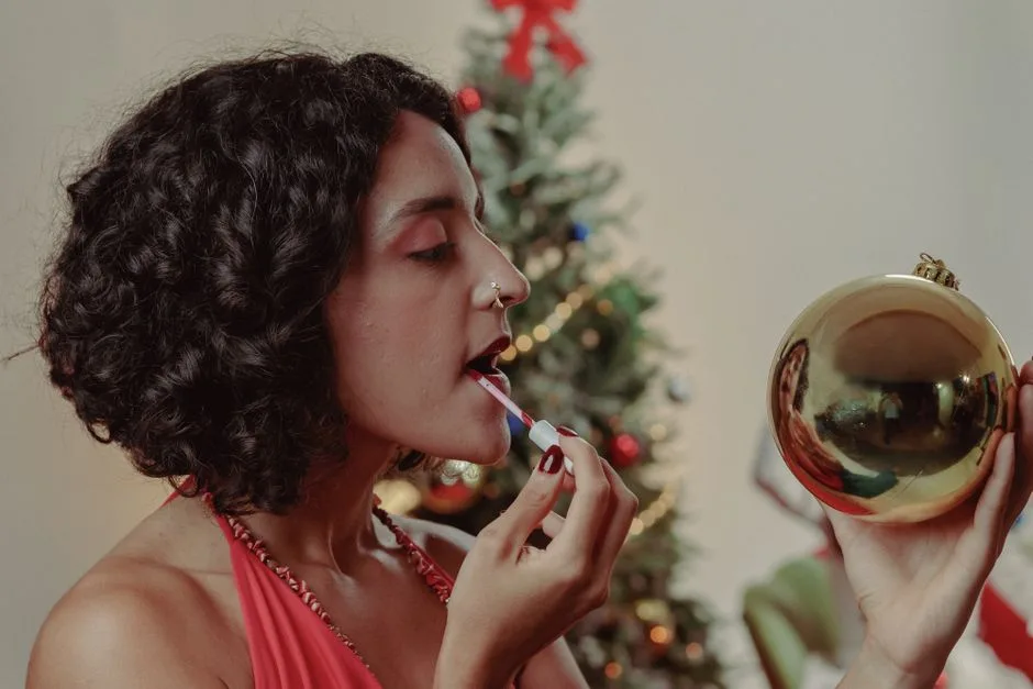 A woman in a red dress applies lipstick using a bauble as a mirror near a decorated Christmas tree.